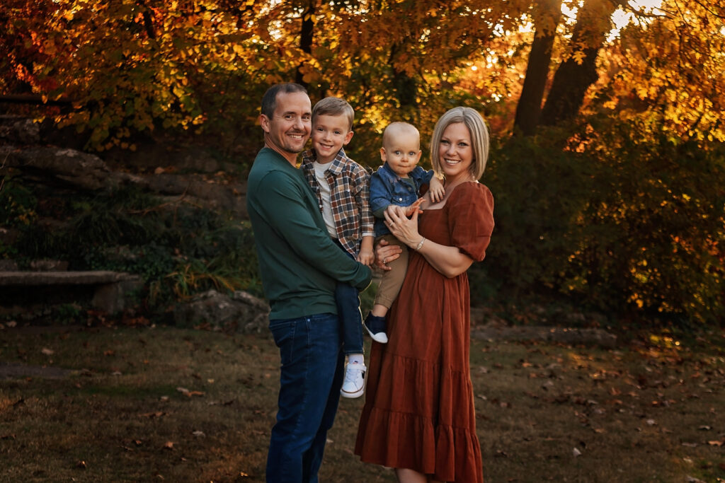Family of four smiling outdoors during a fall photography session surrounded by golden leaves — taken by Shelly Sims Photography in Owasso.