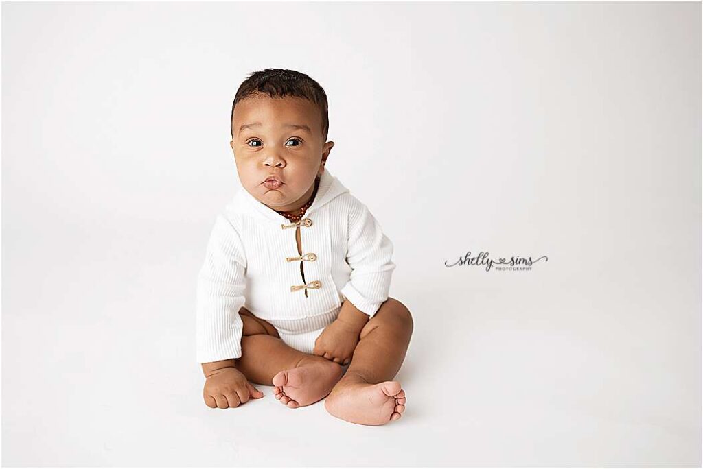 Baby sitting on a white backdrop in a cozy white outfit, looking curiously at the camera — captured during a milestone session by Shelly Sims Photography in Owasso.