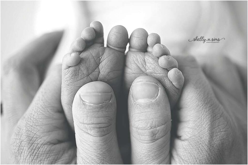 Black and white close-up of a newborn’s tiny feet cradled gently in a parent’s hands — captured during a newborn and milestone photography session in Owasso by Shelly Sims Photography.