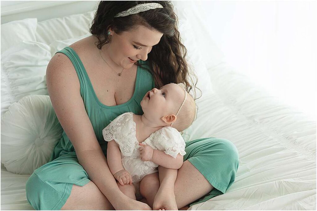 Mother in a soft green dress sitting on a white bed, smiling lovingly at her baby — part of a milestone session with Shelly Sims Photography in Owasso.