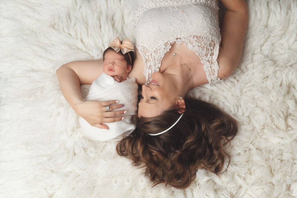 Mother lying on a soft white blanket, smiling as she cuddles her newborn baby wrapped in a white swaddle with a peach bow — captured during a newborn and milestone photography session in Owasso by Shelly Sims Photography.
